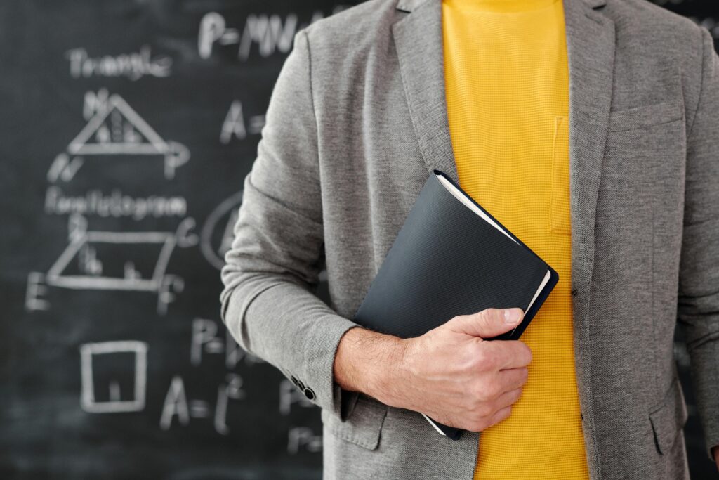 pexels-photo-5212360-5212360 Close-up of a teacher with a notebook standing in front of a blackboard with equations.